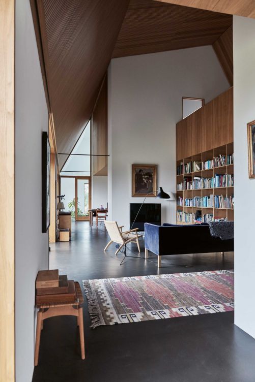Das Wohnzimmer wird von einem beeindruckenden Bücherregal bestimmt, das ebenfalls aus Eichenholz gefertigt wurde. Die weiss getünchten Wände und das Holz sollen an die traditionellen Bauernhäuser mit freigelegten Balken erinnern. Die Einrichtung ist schlicht und gemütlich gehalten mit dänischen Klassikern und persönlichen Erbstücken. Das Sofa ist von Børge Mogensen; die Stühle sind von Hans Wegner. Das Gemälde über dem Fernseher stammt von Ejnar Nielsen und ist ein Erbstück von Stines Grossmutter. Stehleuchte «Costanza» von Paolo Rizzatto für Luceplan; Teppich: Märtha Maas Fjetterstrøm; Stehleuchte «Mantis» von Bernard Schottlander, 1951.
