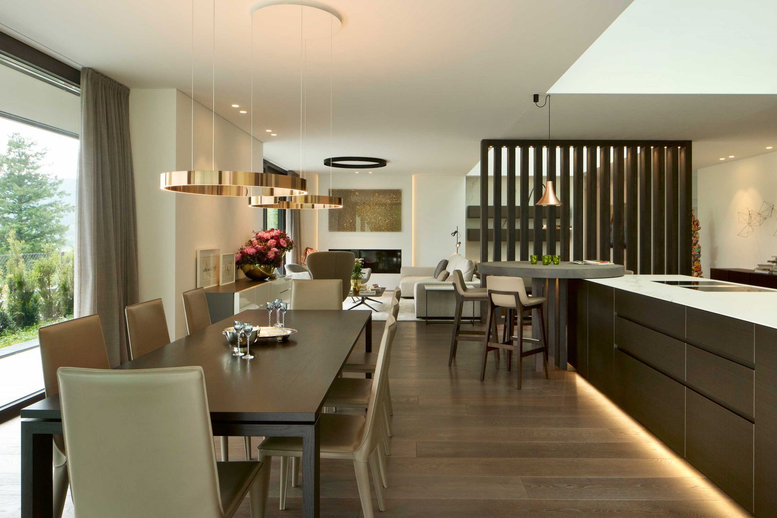 Dunkelbraune Holzstaketen trennen den Wohnbereich von der Küche und verbergen zeitgleich zwei tragende Stützen. In Küche und Essbereich kombinierten André und Irène Roesch-Zwyssig eigene Entwürfe aus der Trendline Designmanufaktur und Möbel des italienischen Herstellers Poliform. Pendelleuchten: Occhio «Mito».