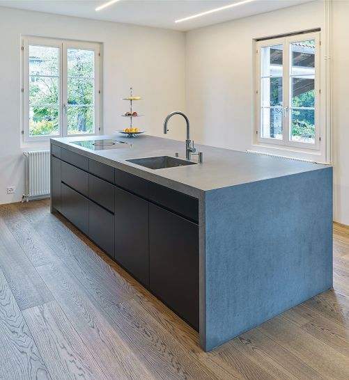 Geradlinig und ästhetisch präsentiert sich diese Küche in einer Kombination aus anthrazitfarbenem Hochleistungsbeton und schwarzen, spritzlackierten Fronten. Das Waschbecken aus Beton ist fugenlos in die Abdeckung integriert, das Kochfeld mit Muldenlüfter flächenbündig eingebaut. Oesch Innenausbau.